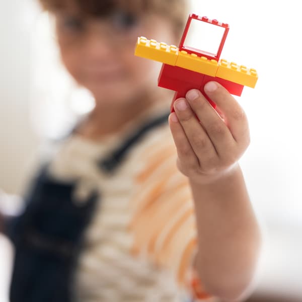 A child stands and holds up a piece of a toy house made out of parts from the BYGGLEK 201-piece LEGO® brick set.