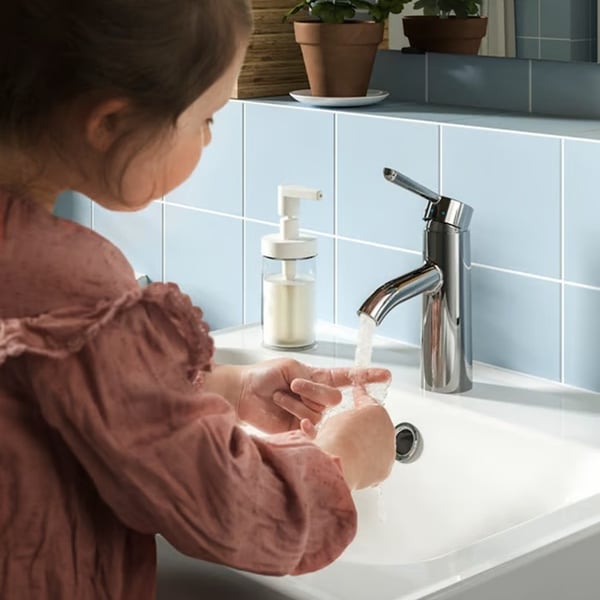A child washes their hands at a bathroom sink with a soap dispenser and chrome tap nearby.