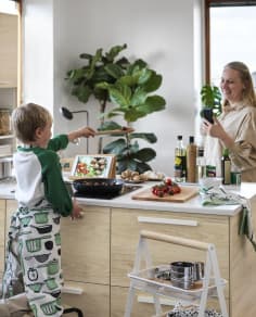 A child wearing a kitchen apron cooking in a pan while a smiling woman takes a photo of him with her phone in a bright kitchen.