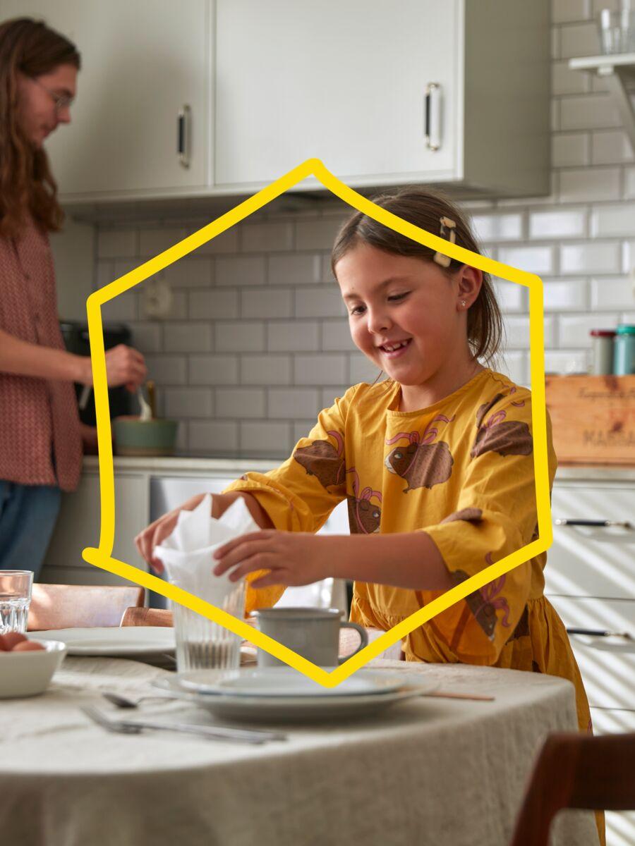 A child wearing a yellow dress places a napkin in a glass on a table, while another person in a bright kitchen mixes ingredients in a bowl on white-tiled countertops.