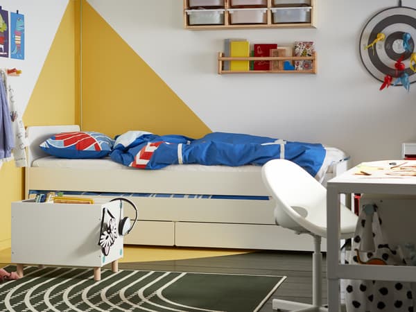 A children’s bedroom with graphic white and yellow walls, a white bed with storage underneath, blue and red bed linen.