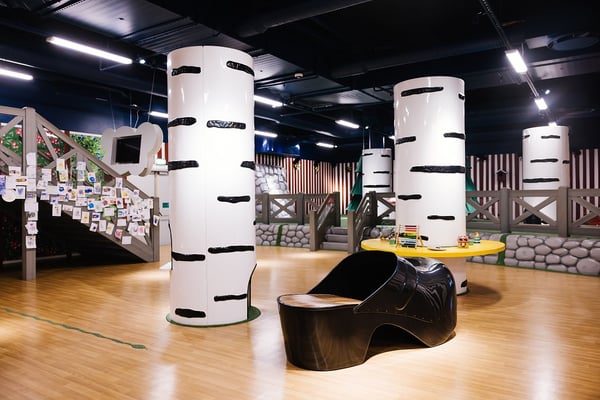 A children’s indoor play area with a forest-themed design, featuring large white tree-like pillars, a yellow activity table with toys, a black play structure shaped like a horse, and a staircase decorated with kids’ drawings.