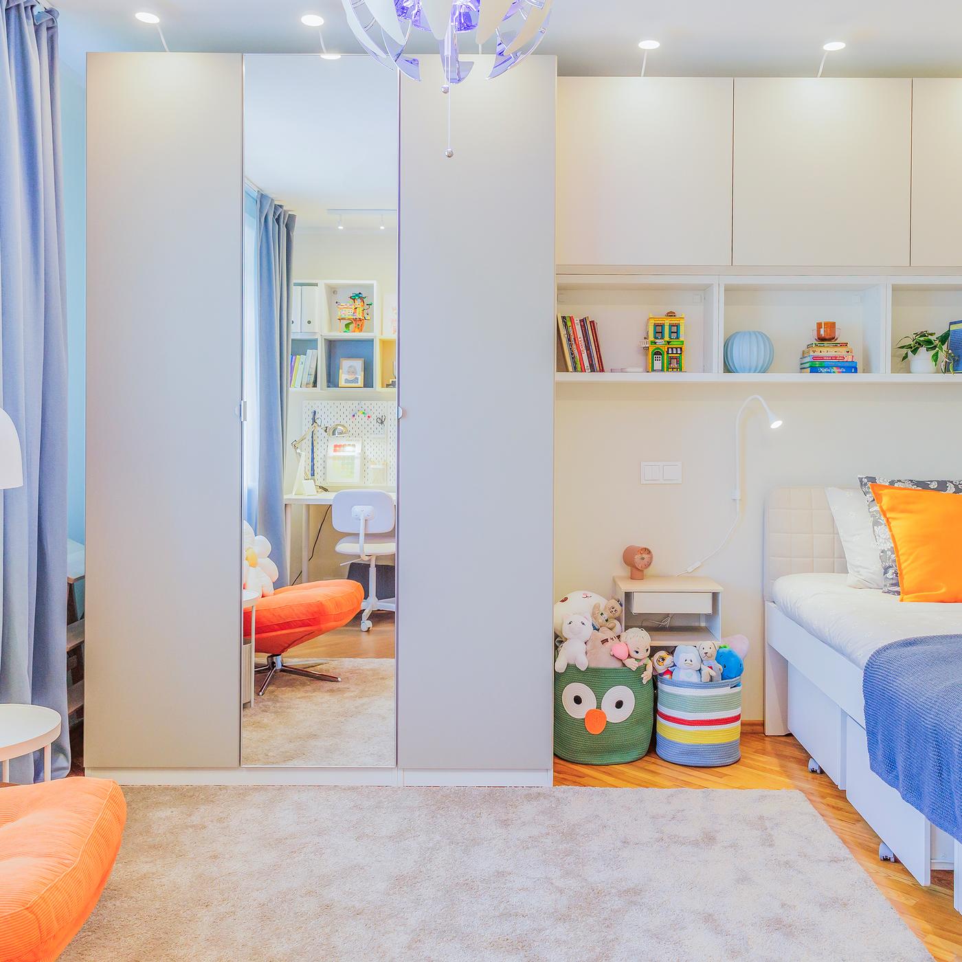 A children’s room after a makeover, featuring a bed with colourful bedding in shades of blue and orange. Open shelves and a mirrored wardrobe line the wall, with natural light streaming in from the window.
