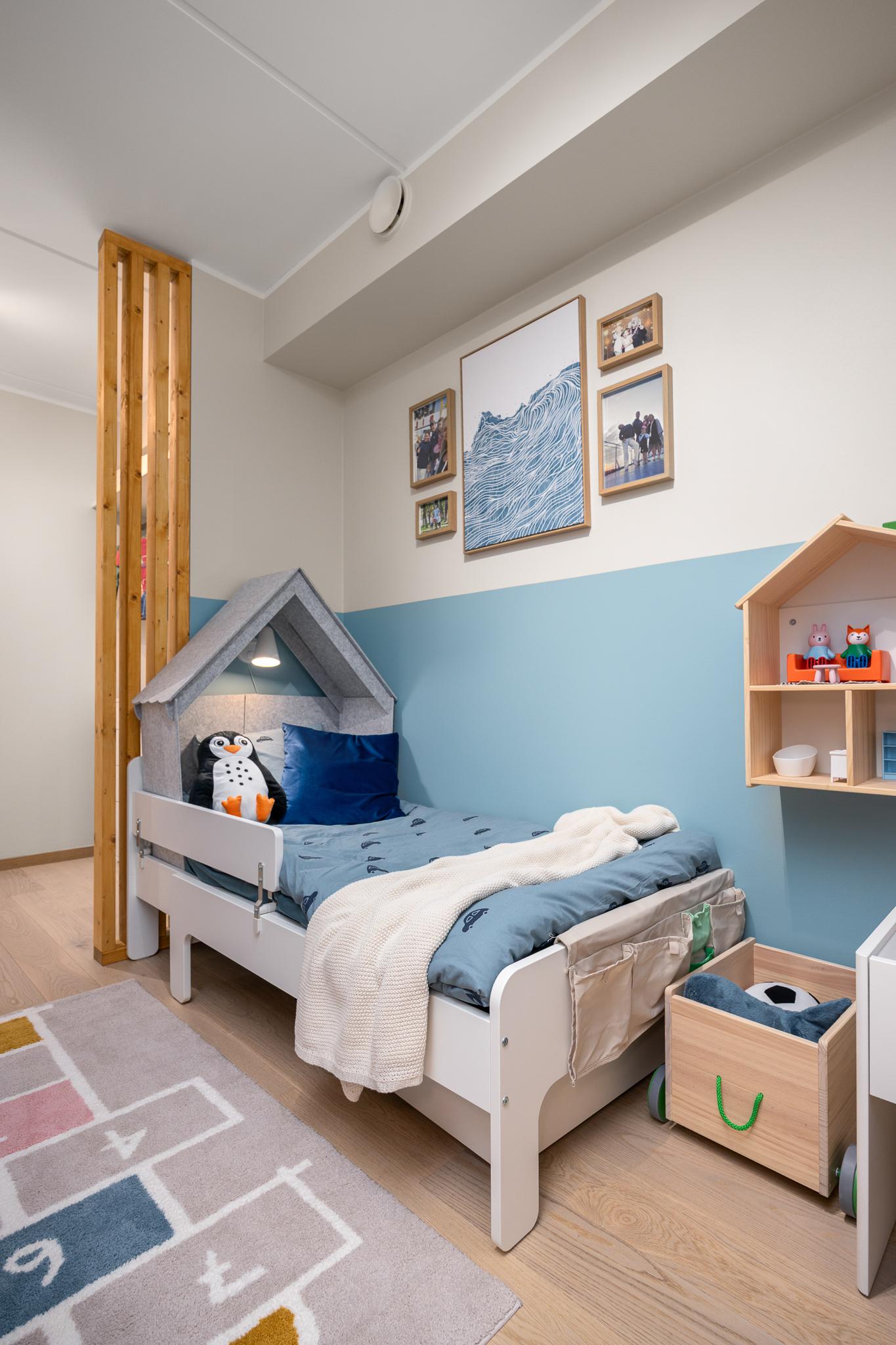 A children’s room in light tones with a house-shaped bed. A soft rug covers part of the floor, and toys are placed in the corner. Wall shelves hold books and small items.