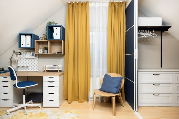 A children’s room with a study area next to a wardrobe zone, featuring a desk, chair, wall shelves, yellow curtains, and sliding doors separating the spaces.