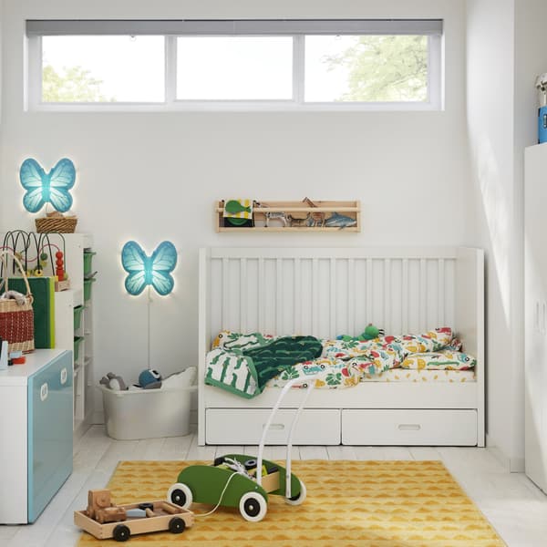 A children’s room with a white cot, a yellow rug, a green toddle truck and two blue butterfly-shaped wall lamps.