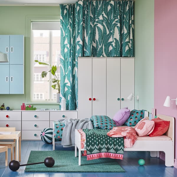 A children’s room with white wardrobes and chests of drawers, two extendable bed frames, a green rug and blue wall cabinets.