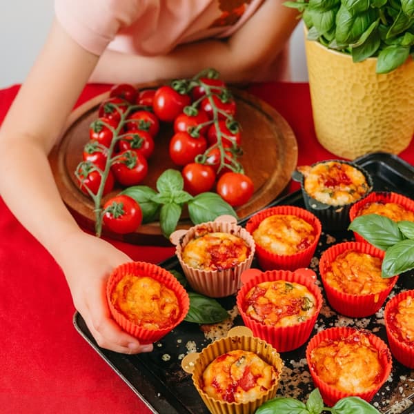 A child's hand holding a salty muffin in a silicone mold. Several freshly baked muffins and basil leaves are arranged on a pan, cherry tomatoes in clusters on a plate next to them.