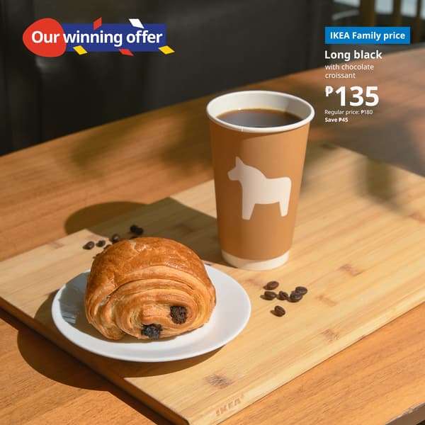 A chocolate croissant on a white plate and a paper cup of coffee with a horse logo sit on a wooden table. Price details in IKEA branding are visible.