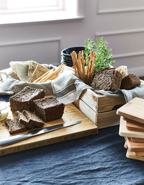 A chopping board and wooden crate filled will bread, crackers and bread sticks on a table.