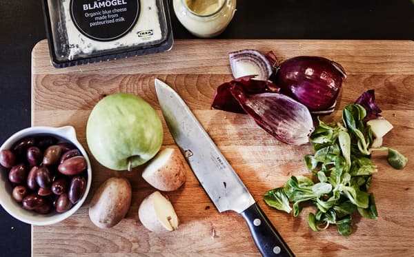 A chopping board with olives, an apple, potatoes, red onion, greens and a knife