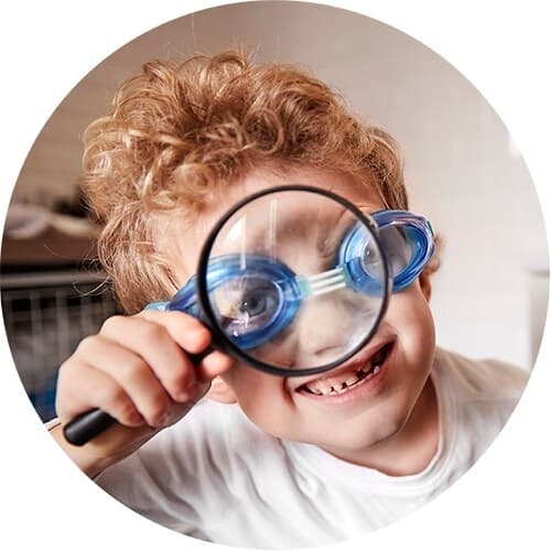 A circular image of a child wearing goggles holding a magnifying glass to his face.