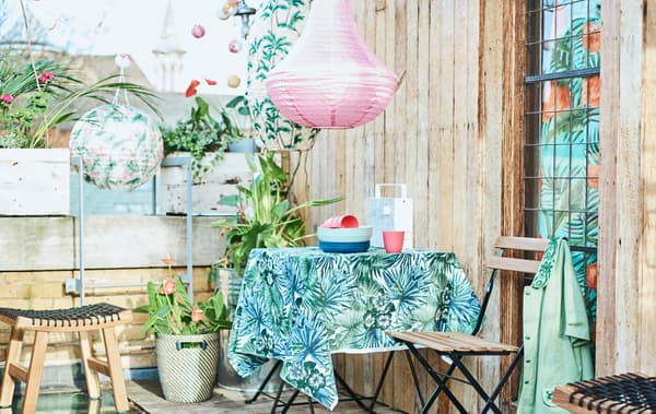 A city roof terrace with a patio table covered with leaf-print fabric, folding chairs, potted plants and coloured lanterns.