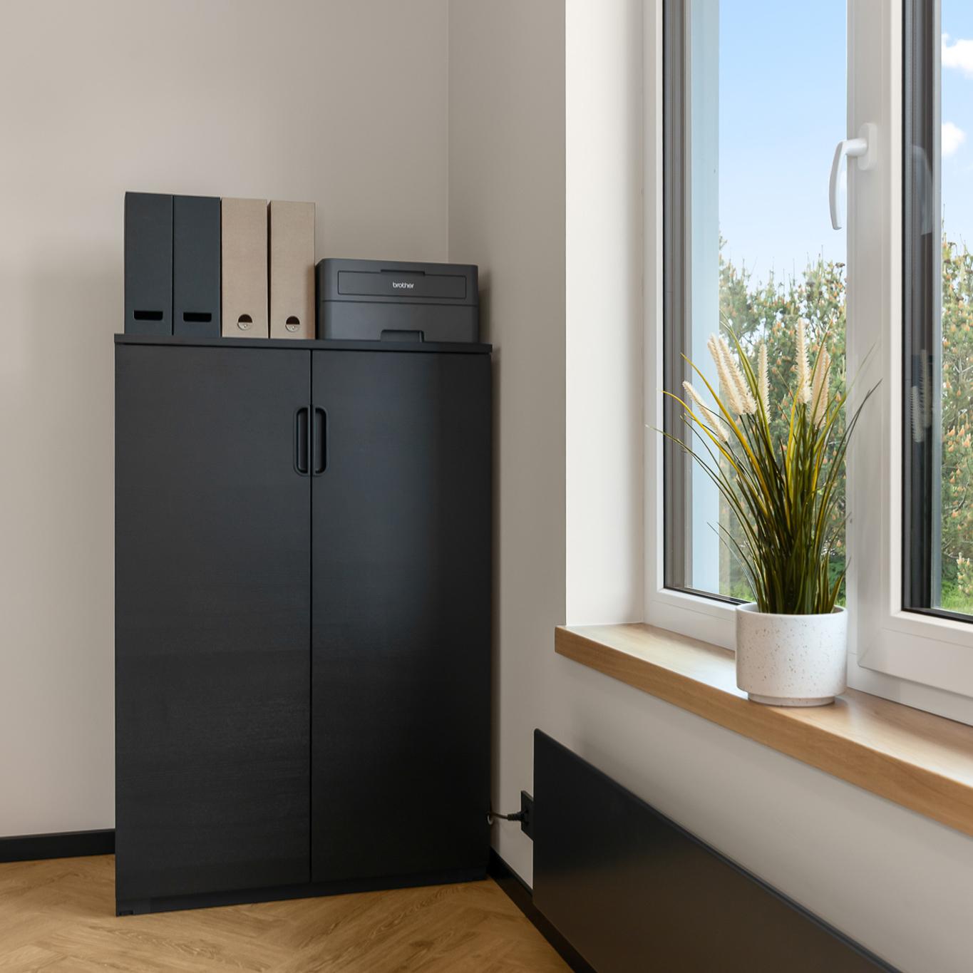 A clean office corner featuring a tall black storage cabinet topped with neatly arranged magazine files and a compact printer, set against a light wall and wood flooring next to a large window.