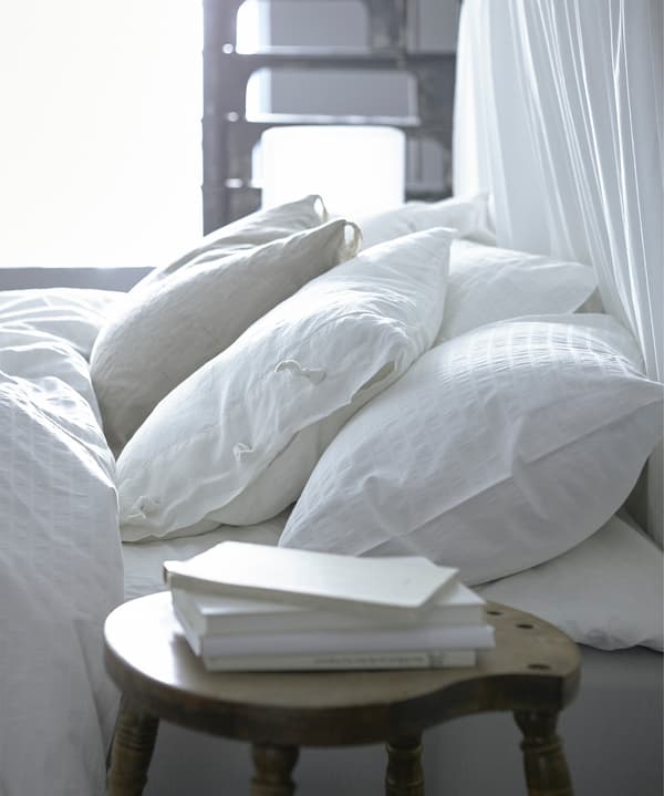 A close-up image of white pillows lying on a bed and with some white books on the bedside table.