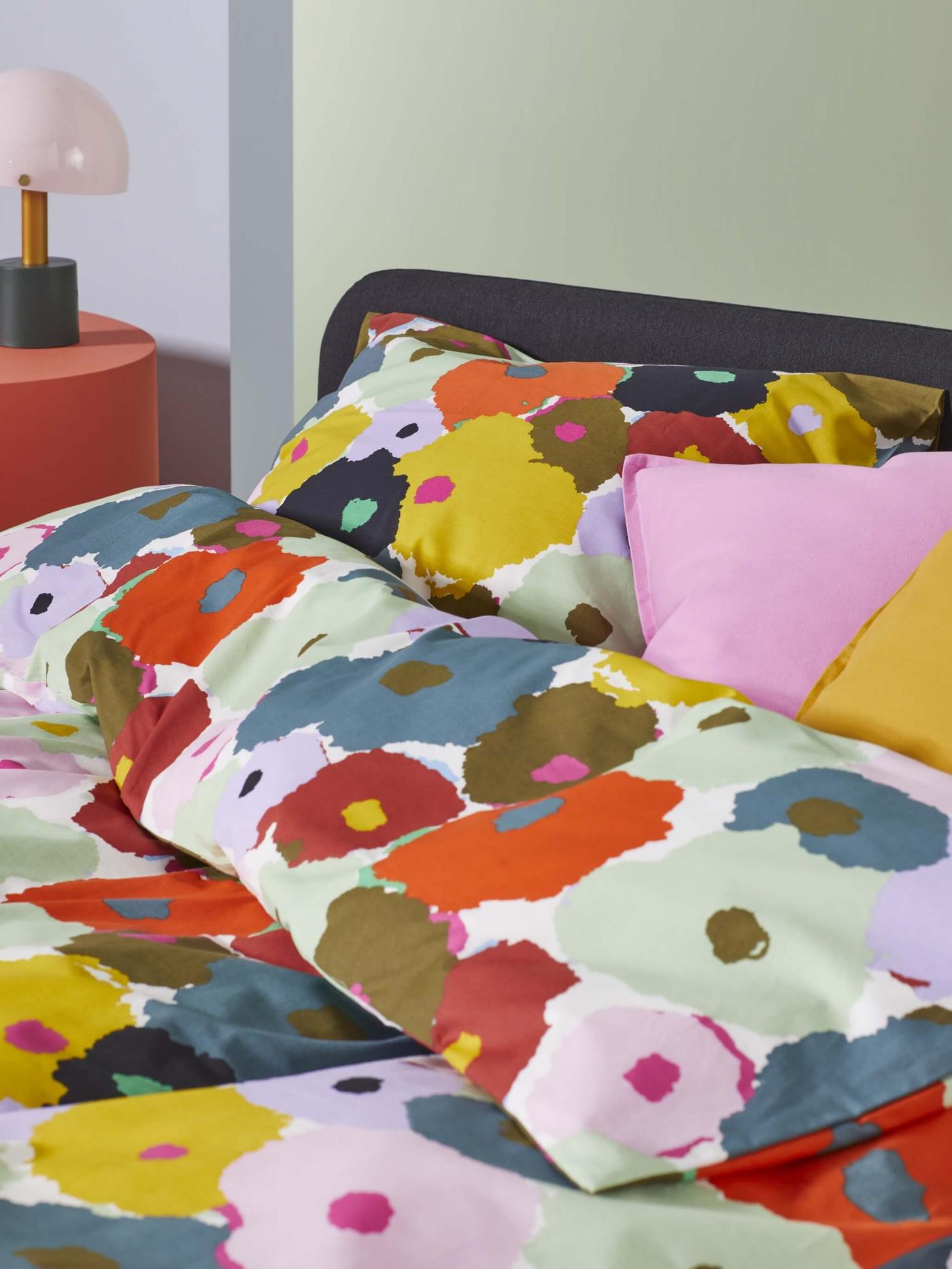 A close-up of a bed with floral colorful bedding. The duvet cover and pillows feature a vibrant, abstract pattern with large splashes of yellow, red, green, brown, and black.