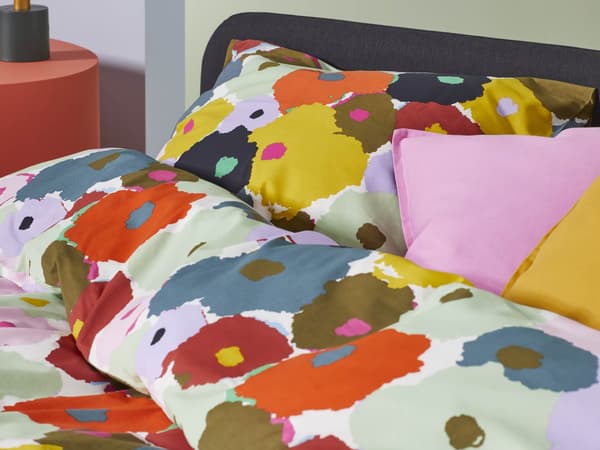 A close-up of a bed with floral colorful bedding. The duvet cover and pillows feature a vibrant, abstract pattern with large splashes of yellow, red, green, brown, and black.