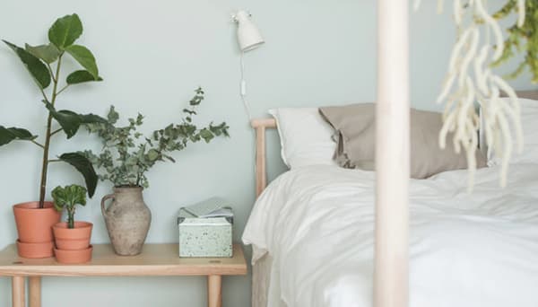 A close-up of a bed with lightly colored, natural textiles against a light green wall and a low ash bedside table with plants and a storage box