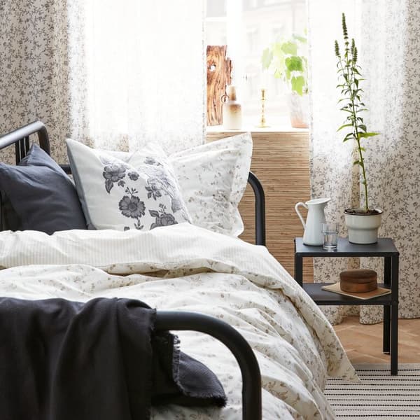 A close up of a bed with STRANDFÄRNE and PRAKTBRÄCKA floral print textiles, ODDRUN cotton throw and a VIGDIS cushion.