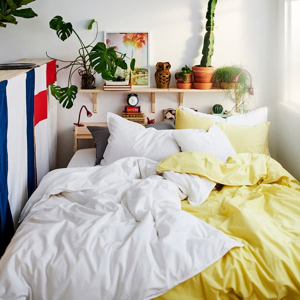 A close up of a bed with white and yellow bedding, a shelf of plants on the wall above.