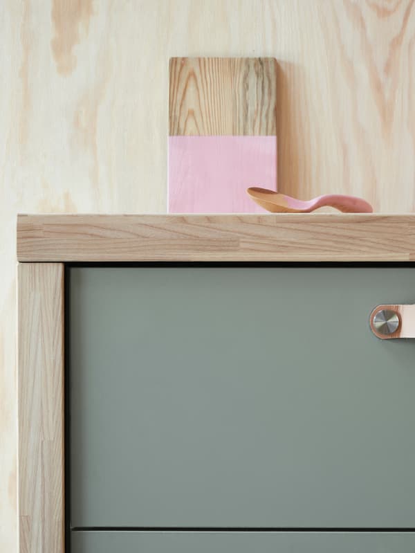 A close-up of a cabinet with a grey-green drawer front against a wooden wall. A half-painted piece of wood is on top.
