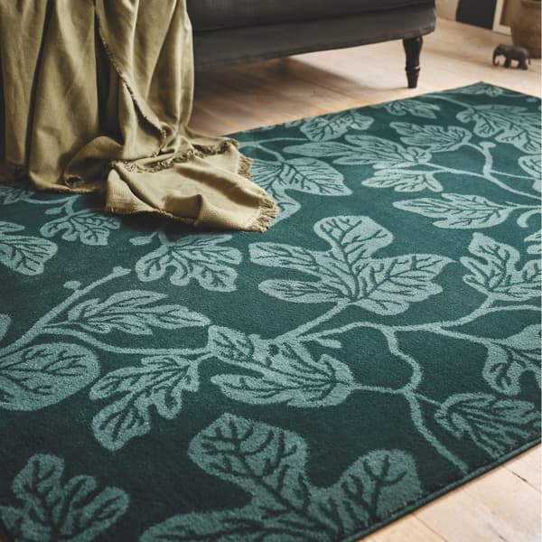 A close-up of a dark green HILDIGARD rug with a fig leaf pattern, a sofa and olive throw can be seen on the side.