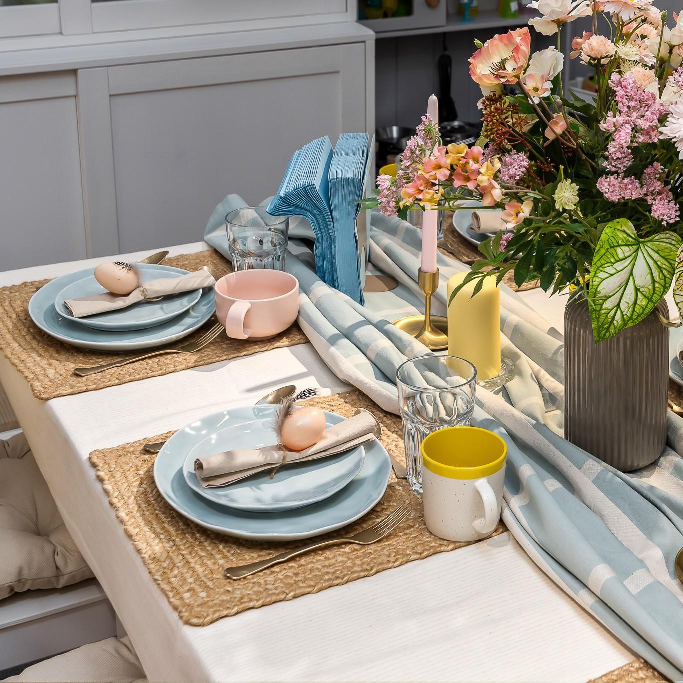 A close-up of a Easter-themed table setting with pastel blue plates, woven placemats, a light blue runner, and a vase of colourful flowers.