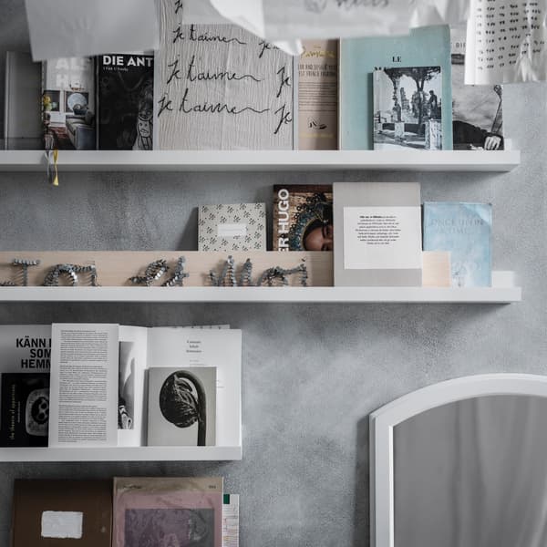 A close up of a grey wall with white wall shelves holding books, papers and decorative images.