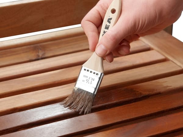 A close-up of a hand holding a paintbrush applying a clear finish to a wooden surface. The bristles of the brush are in contact with the wood, spreading the liquid evenly. The wooden surface has multiple slats, indicating it may be part of furniture or flooring.
