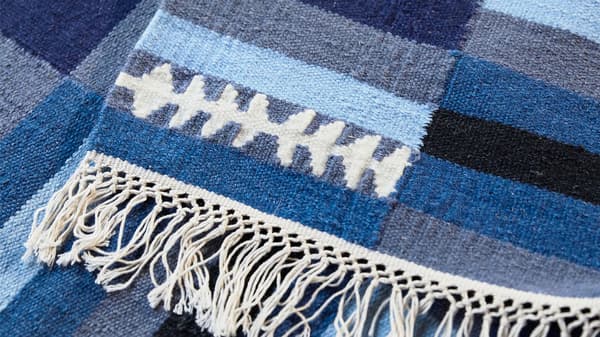 A close-up of a handmade striped TRANGET wool rug in white, black, grey and blue, with white tassels on the edge.