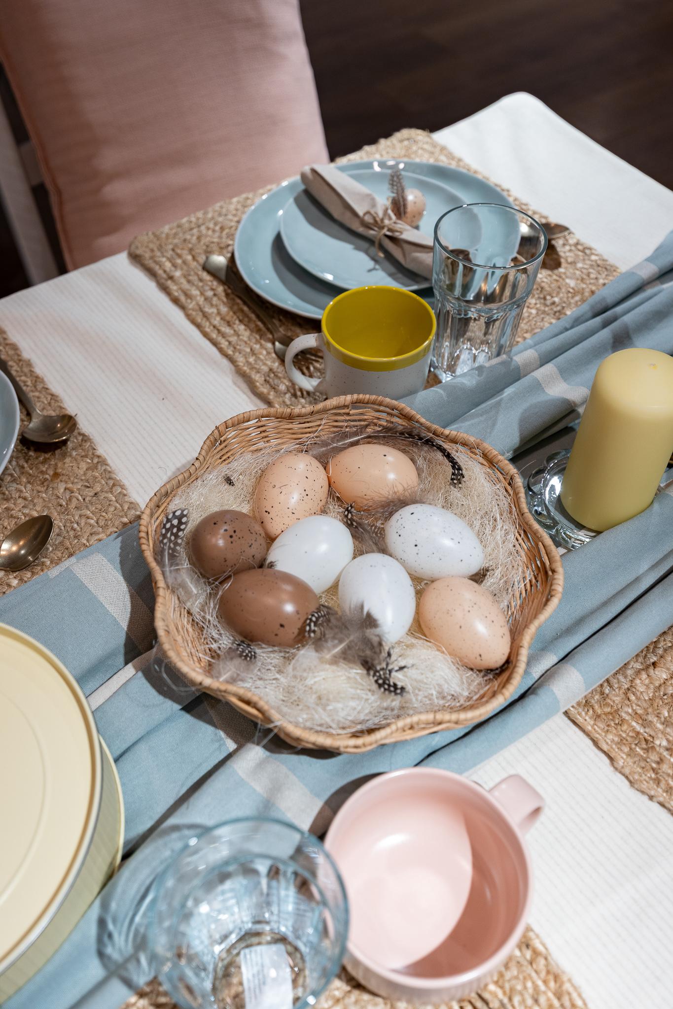 A close-up of a pastel blue place setting with woven placemats, neutral napkins tied with small decorative accents, and soft floral details in the foreground.