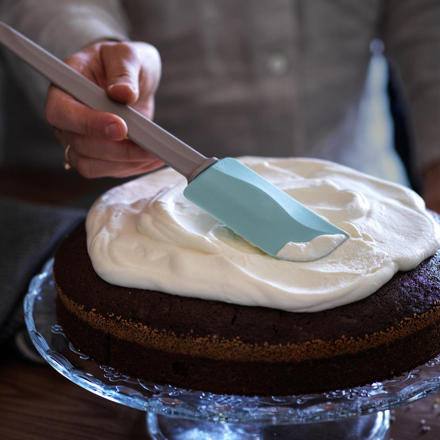 A close-up of a person icing a cake on a stand with a BAKGLAD spatula.