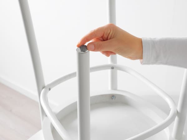 A close-up of a person’s hand placing or adjusting a grey cap on the top end of a white, cylindrical metal leg of a piece of furniture, possibly a chair or table. The background is minimalistic and predominantly white, suggesting an indoor setting with clean and simple design aesthetics.