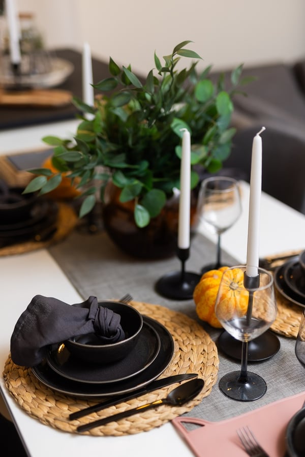 A close-up of a table setting with black plates, glasses, napkins, and decorative pumpkins on a woven placemat.