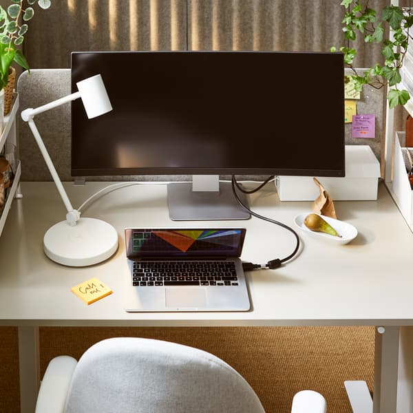 A close-up of a white desk with a white NYMÅNE work lamp, a large computer screen, a laptop and a light grey office chair.