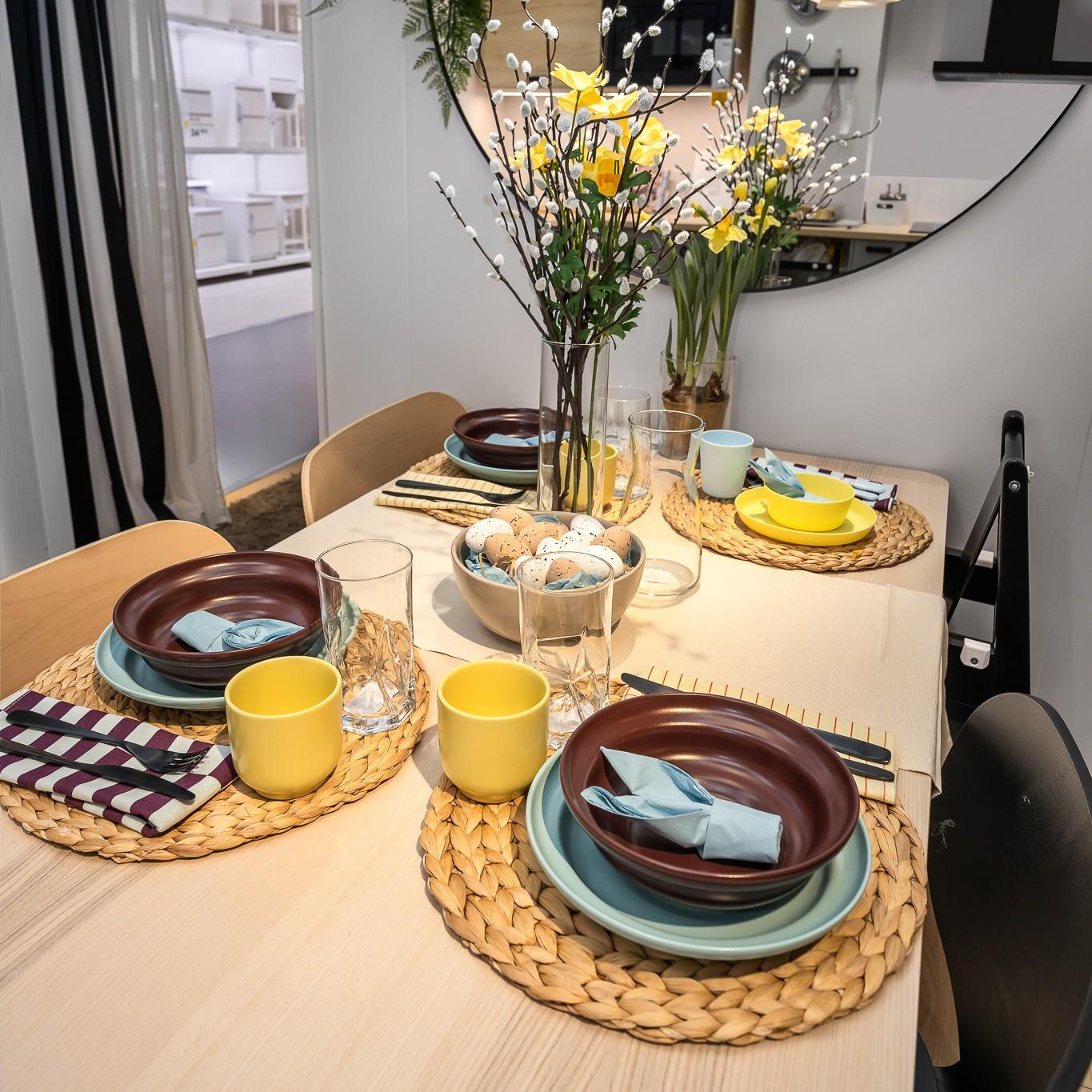 A close-up of an Easter table setting with layered plates, woven placemats, yellow mugs, and folded napkins, decorated with floral arrangements and Easter eggs.