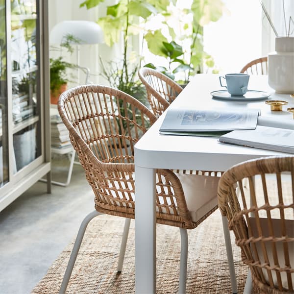 A close-up of an IKEA NILSOVE woven rattan chair next to a white TINGBY table, set with coffee cups and books.