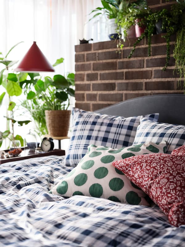 A close-up of an upholstered HAUGA bed, with a checked duvet cover, a spotted green pillow and a flowery red pillow.