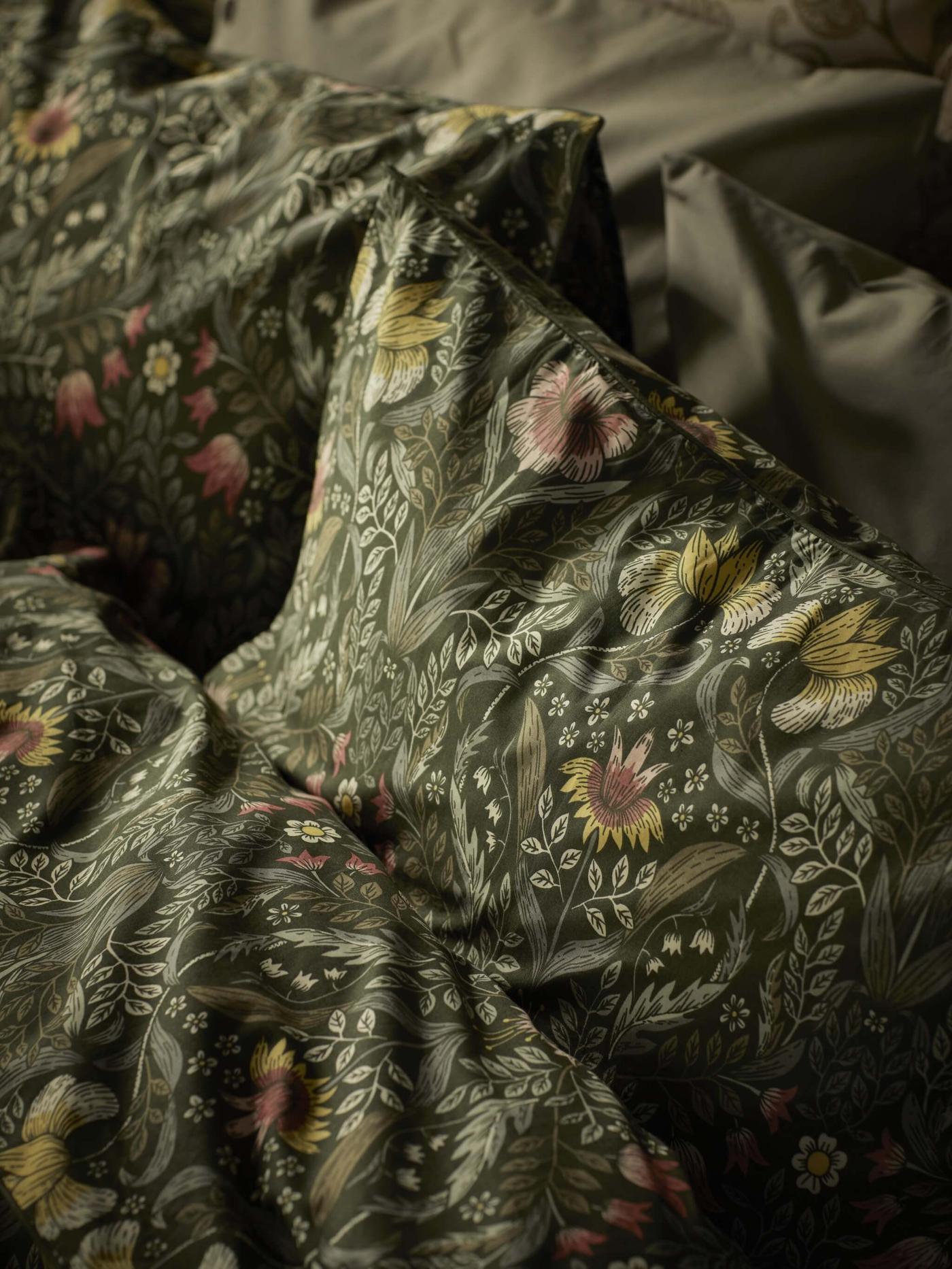 A close-up of bedding with a detailed floral pattern. The pattern features various flowers and leaves in shades of pink, yellow, and green on a dark background.