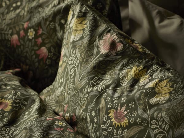 A close-up of bedding with a detailed floral pattern. The pattern features various flowers and leaves in shades of pink, yellow, and green on a dark background.