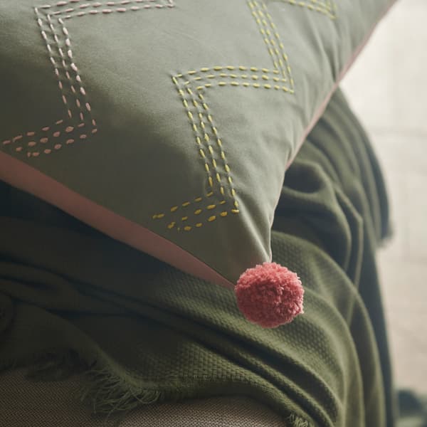 A close-up of MOAKAJSA cushion cover showing pink and yellow embroidery on the green side and a pink pompom in the corner.