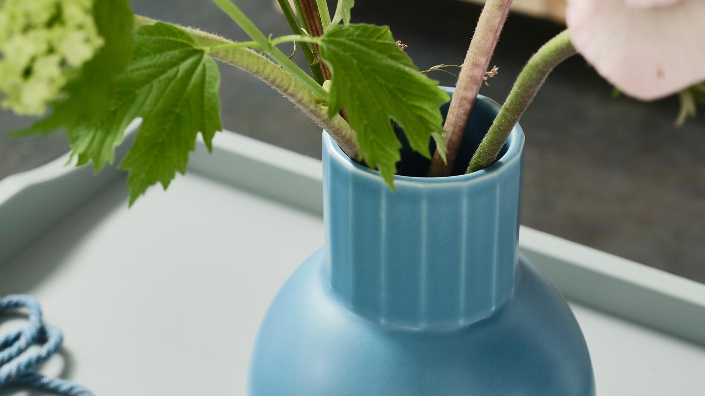 A close-up of OMFÅNG vase in a blue colour standing on a side table holding some green stems of flowers.