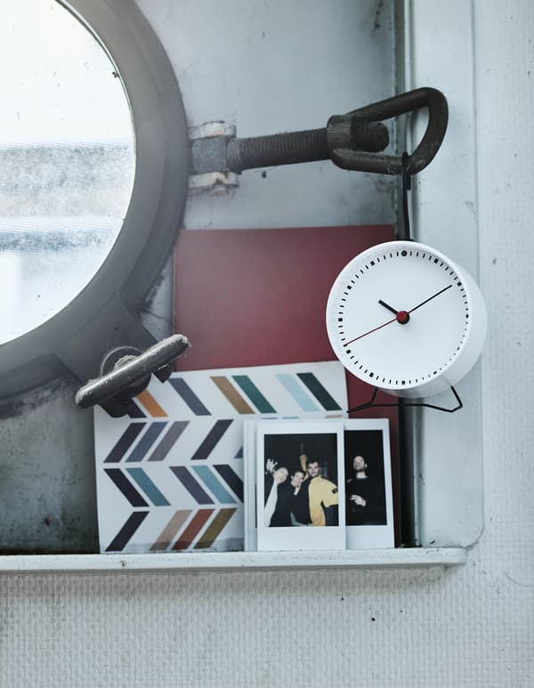 A close-up of pictures and an alarm clock on a small shelf next to a porthole window.