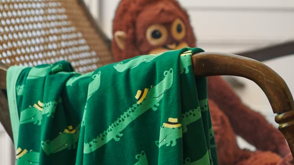 A close-up of RÖRANDE blanket with friendly crocodiles crawling across the green background, placed over a wicker chair.