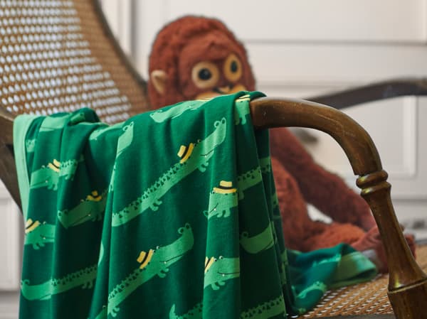 A close-up of RÖRANDE blanket with friendly crocodiles crawling across the green background, placed over a wicker chair.