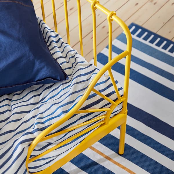 A close-up of the corner of a yellow MINNEN bed frame with blue and white striped bedding and rug.