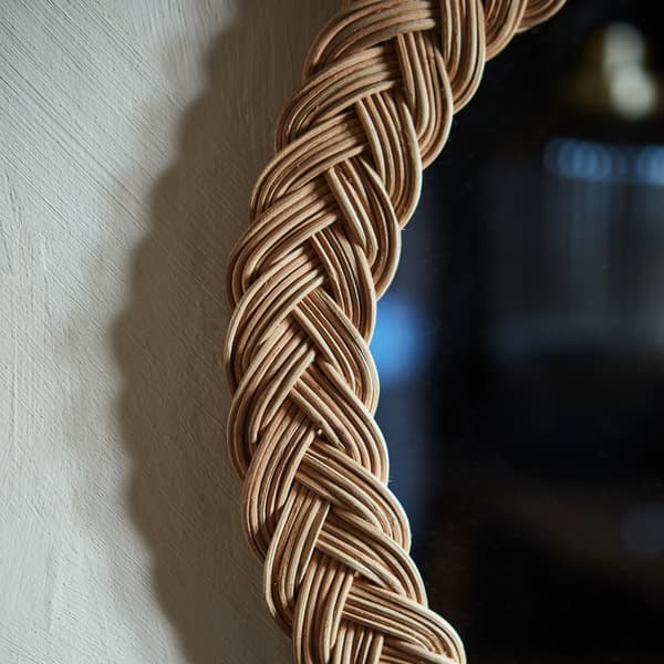 A close-up of the edge of KRISTINELUND mirror made in decoratively woven rattan, hanging on a grey wall.