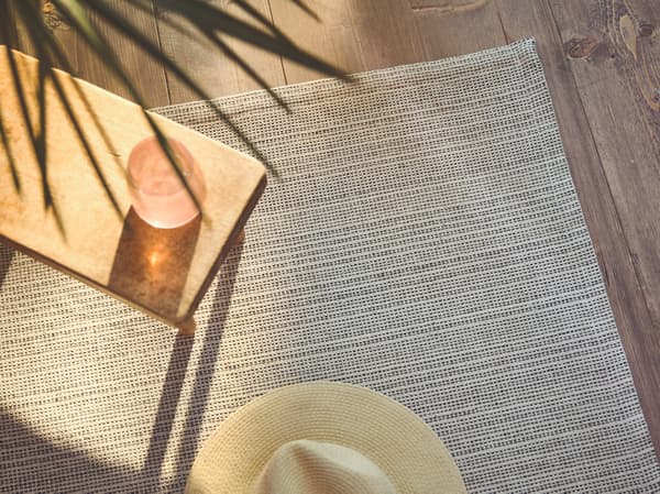 A close-up of the flatwoven TIPHEDE rug with a side table and Panama hat placed on it, while sunlight streams in on one side.