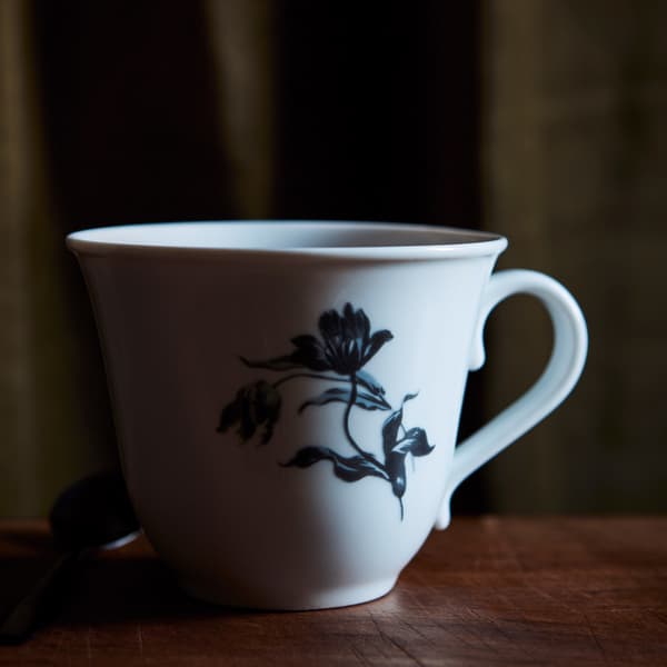 A close-up of UPPLAGA mug with a rustic floral pattern on white placed in a black room with shadows.