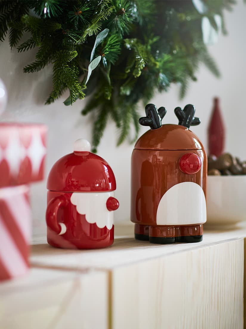 A close-up shot of a red ceramic VINTERFINT Santa bowl with lid beside a brown VINTERFINT reindeer bowl with lid placed on a pine IVAR cabinet.